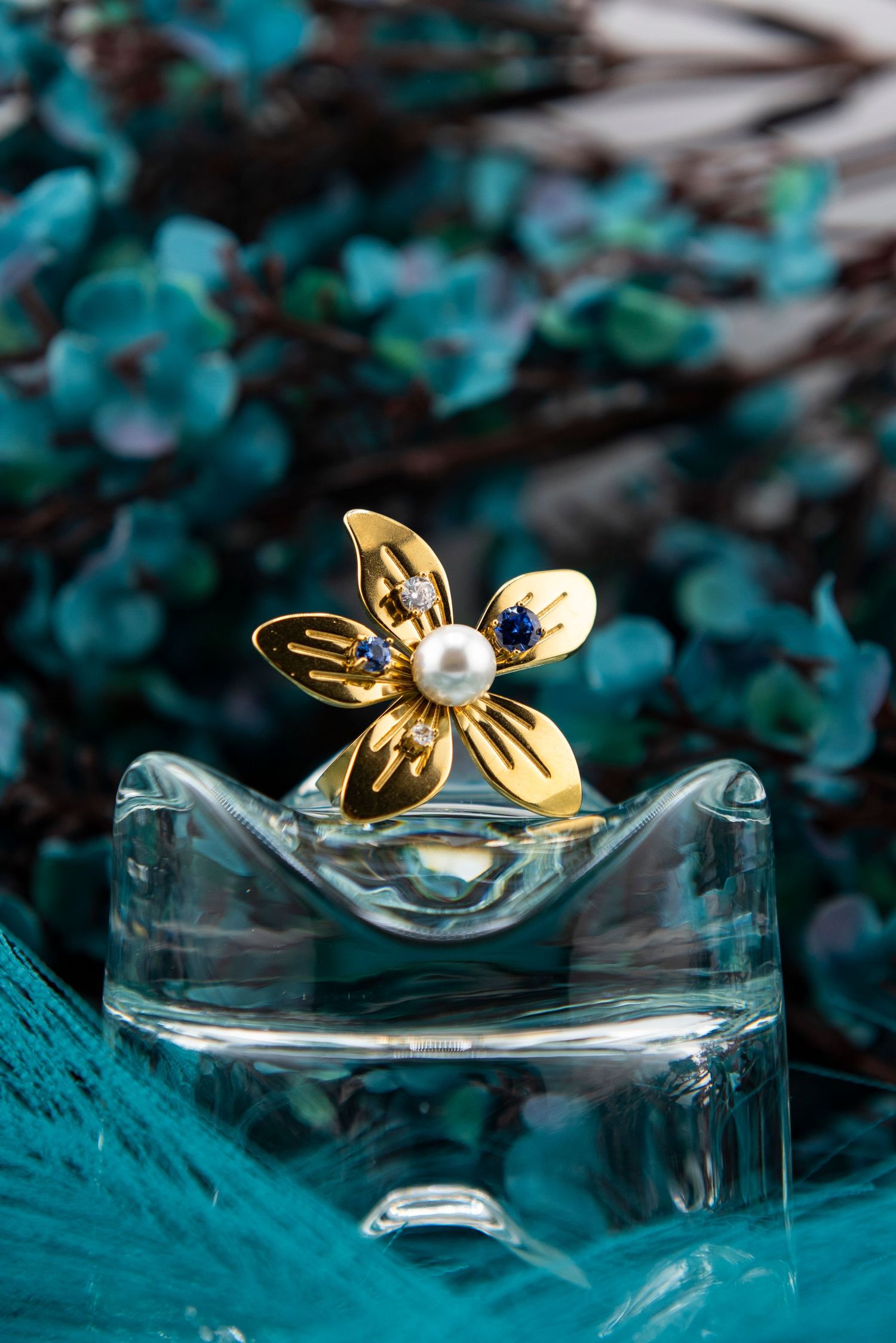 Gold butterfly ring with blue gemstone rests on clear glass surface against turquoise background in macro photography.