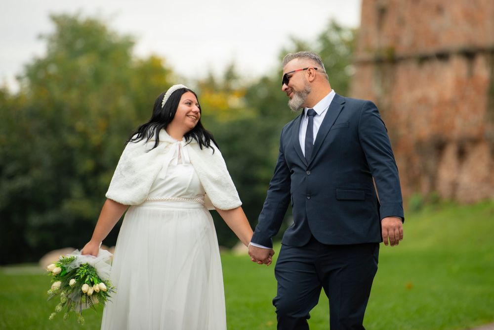 Couple in wedding attire walking together outdoors with green scenery and brick building in background.