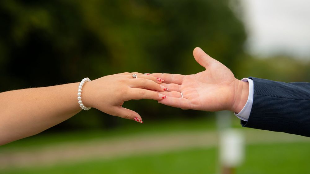 Two hands reaching toward each other against a blurred outdoor background in a romantic gesture.