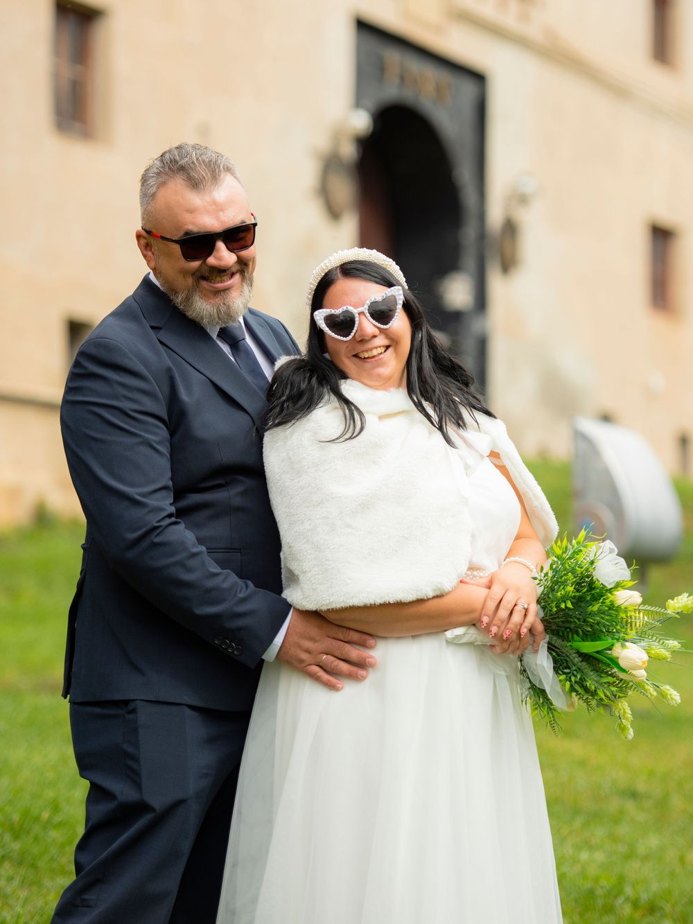 A joyful couple in wedding attire share an intimate moment outdoors on a sunny day near a stone building.