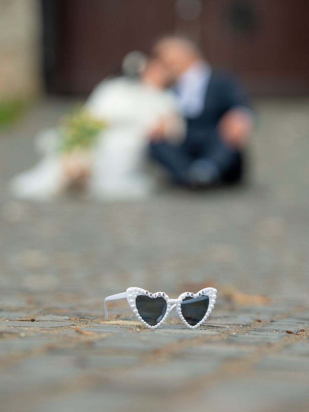 Blurred romantic couple in background with white heart-shaped sunglasses in focus on stone ground.