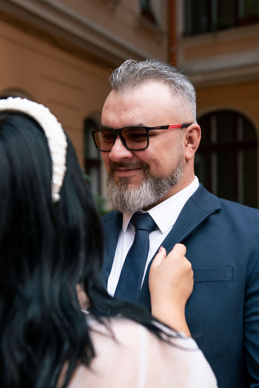 A person wearing glasses and a navy suit smiles warmly while someone adjusts their tie.