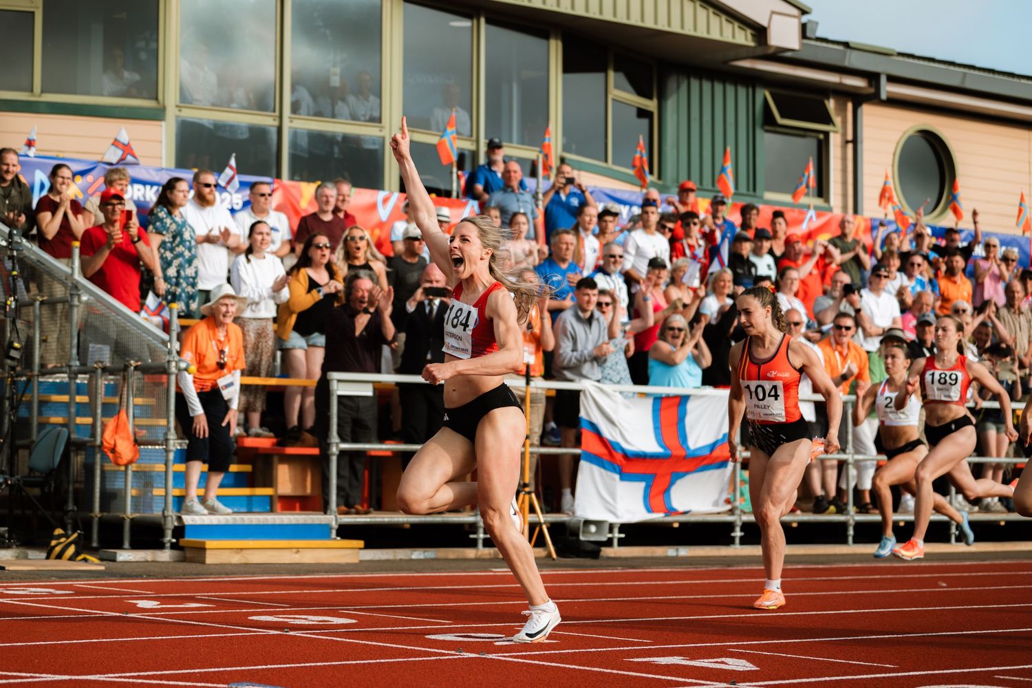 Athletes compete in a track and field relay race with spectators watching from the stands in the background.