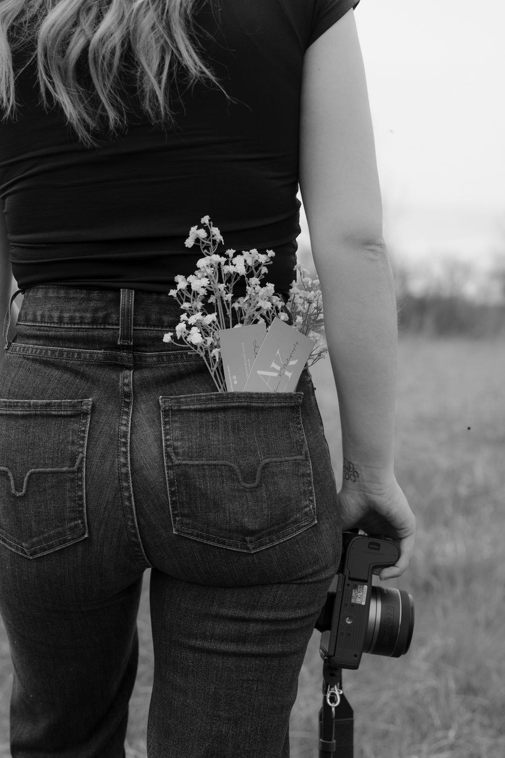 Black and white photo of woman holding camera with wildflowers tucked in back jeans pocket.