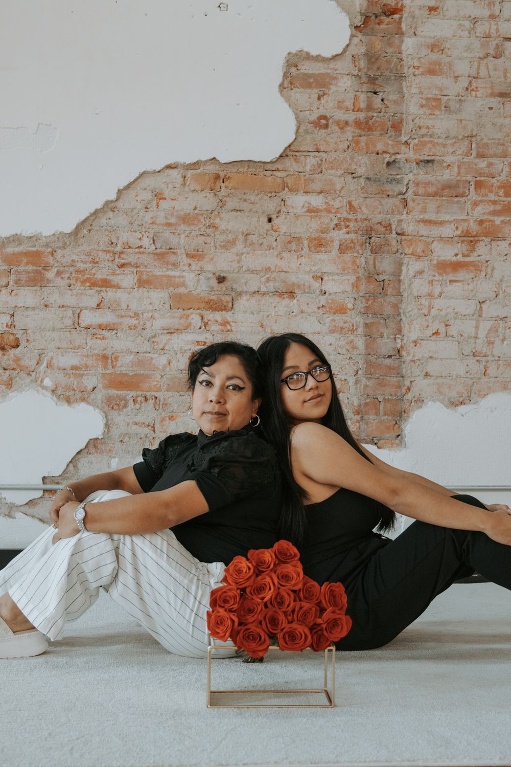 Two women sitting back to back on floor with bouquet of orange roses between them.