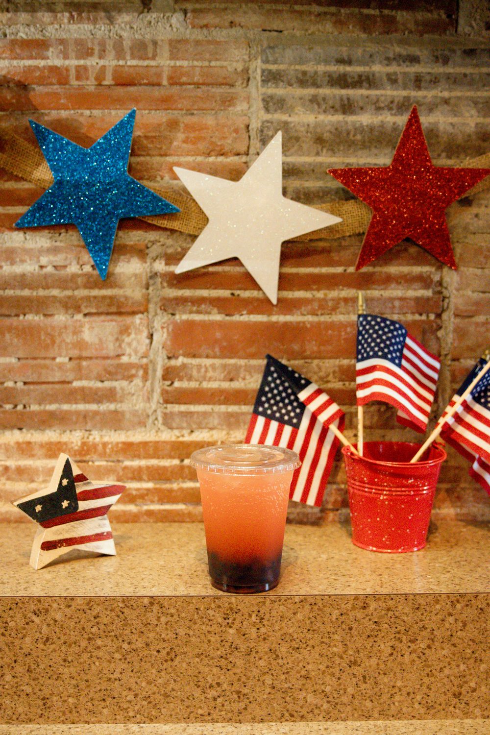 Patriotic Fourth of July display with star decorations, American flags, and layered red drink on cork surface.