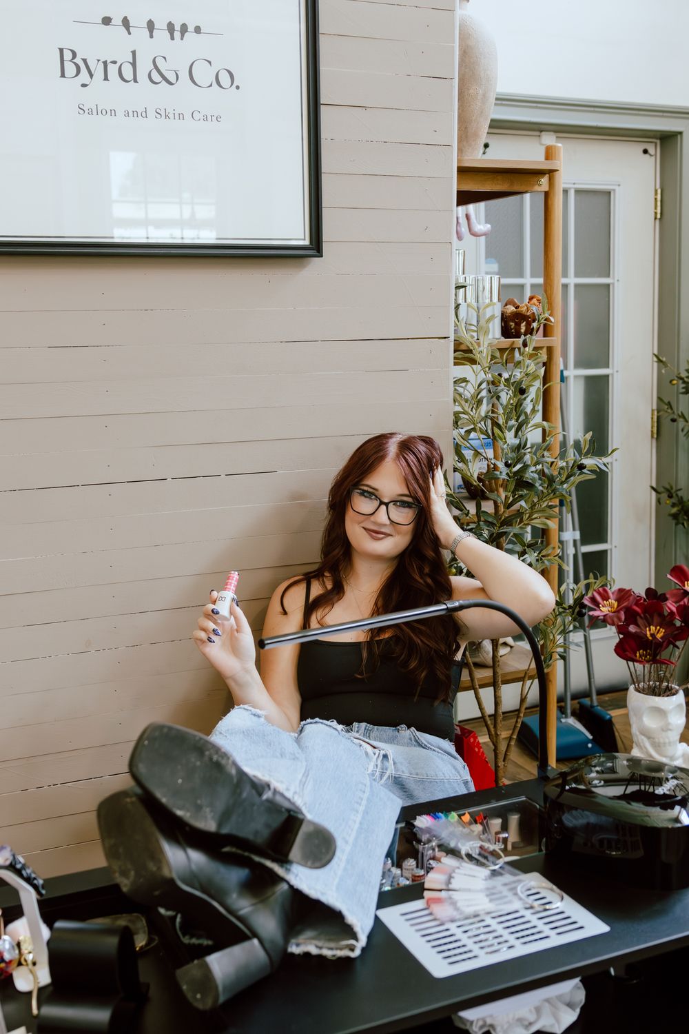 Smiling woman with glasses and long auburn hair sits at a nail station holding polish inside Byrd & Co. Salon and Skin Care.