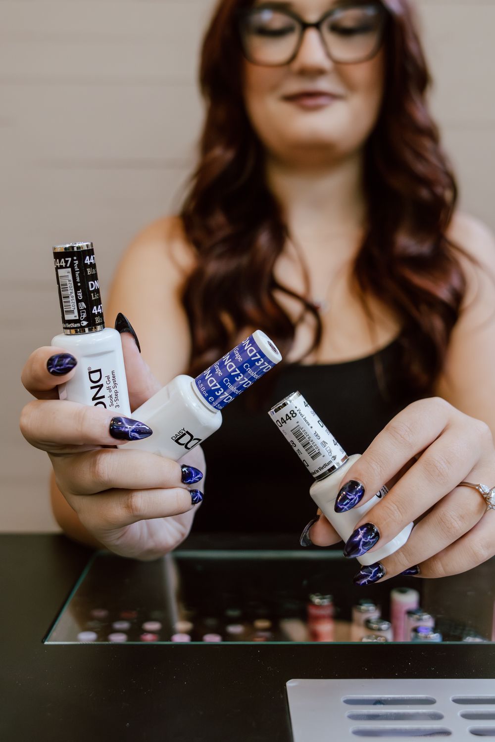 Nail technician with glasses holds three DND gel polish bottles in shades of purple and blue, showing off a matching manicure.