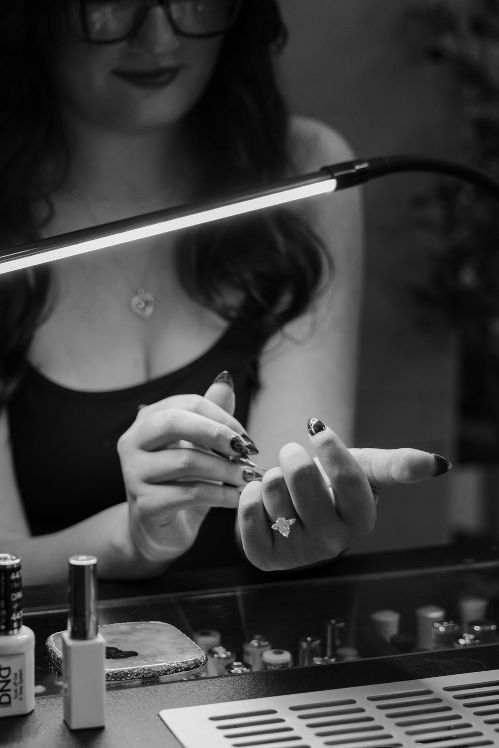 Black and white photo of a nail technician displaying freshly painted nails with a ring, beside a LED lamp and DND gel polish bottles.