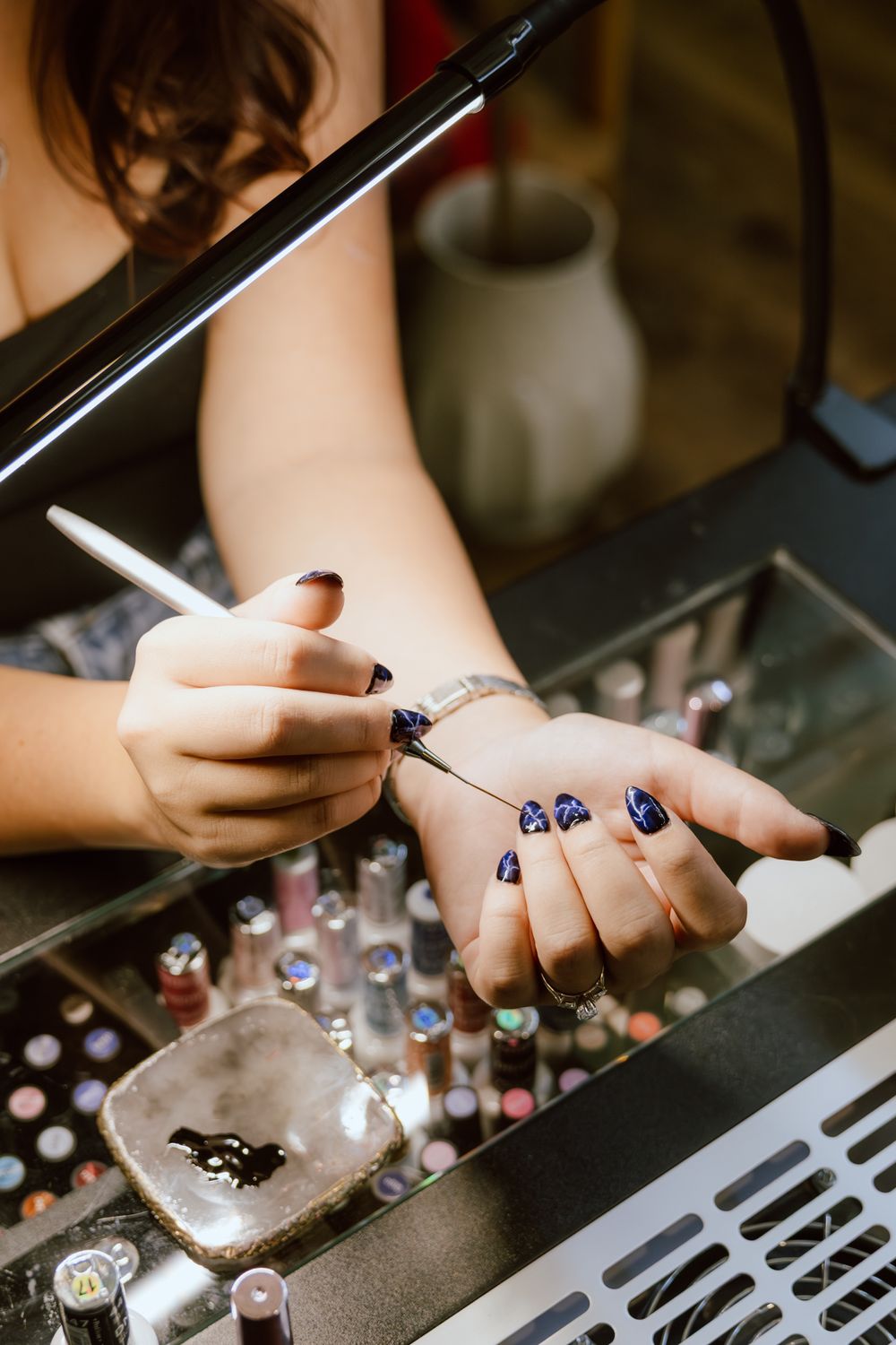Nail technician painting dark navy nail polish with a fine brush, surrounded by rows of polish bottles on a salon display.