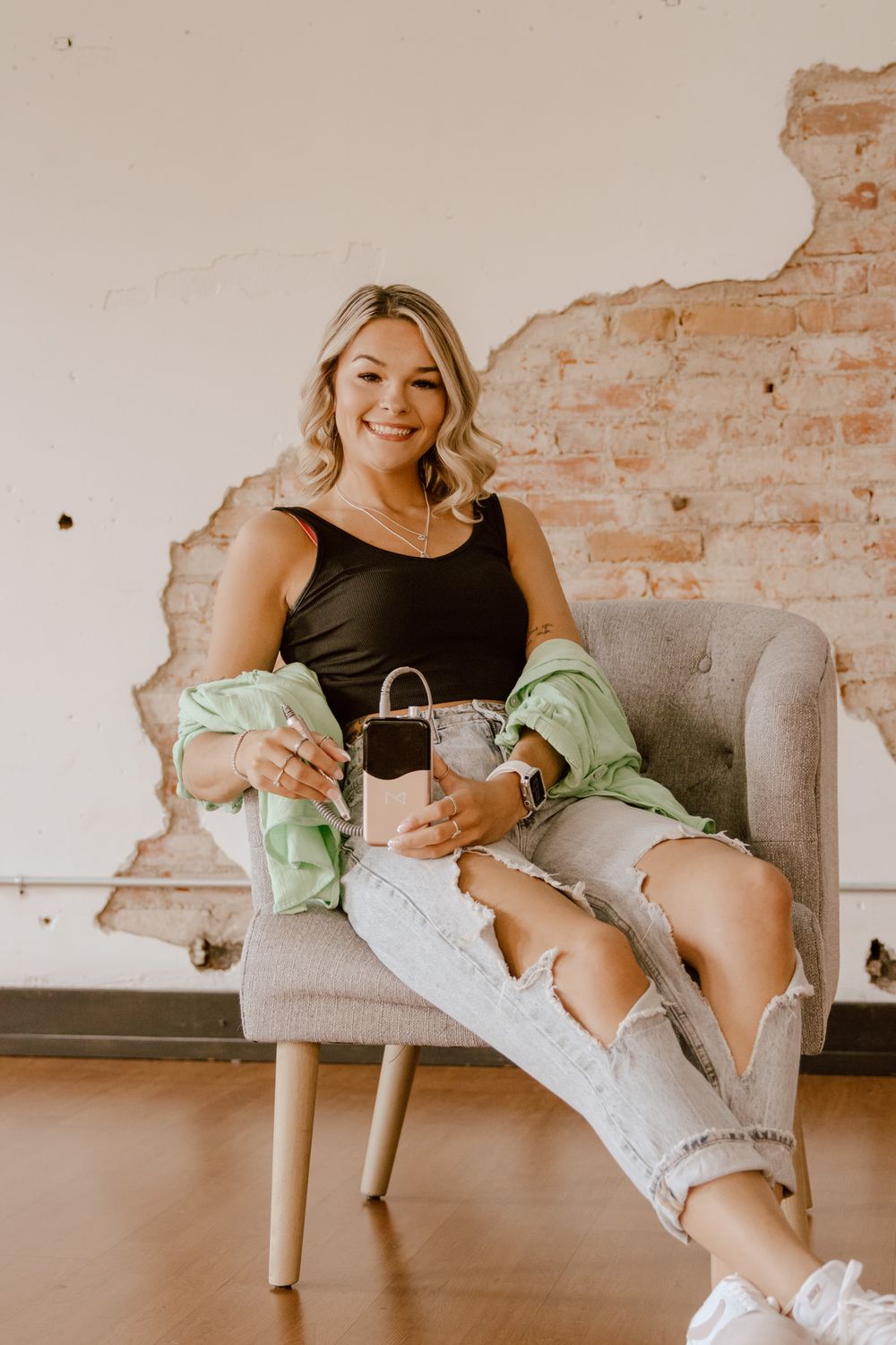 Smiling blonde woman sitting in chair holding phone against exposed brick wall background.