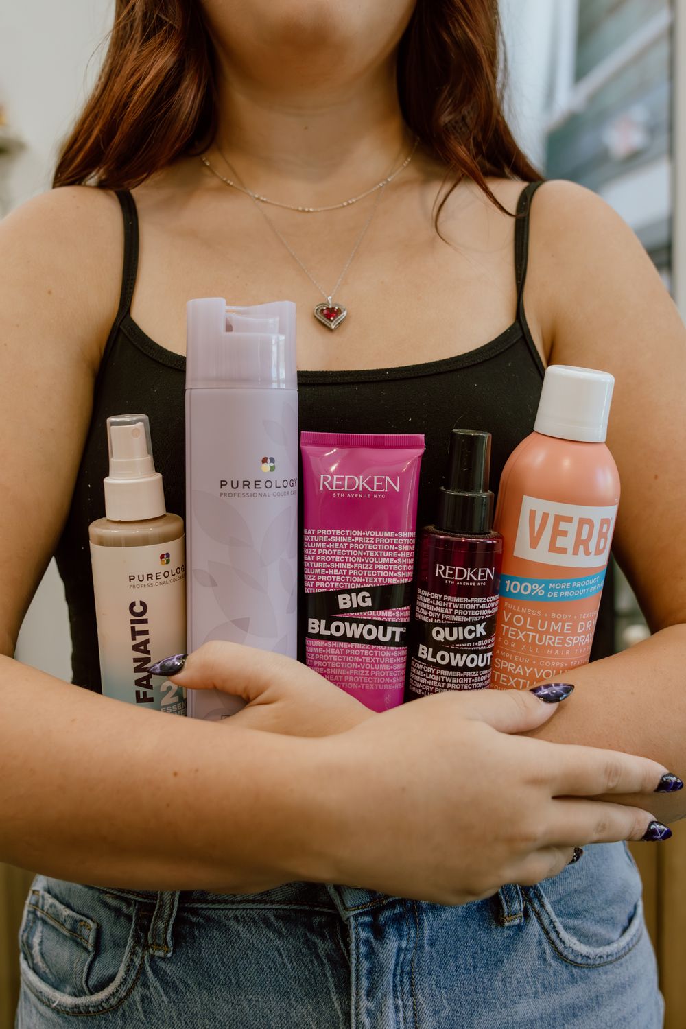 Woman holding Pureology, Redken Big Blowout, Redken Quick Blowout and Verb hair care products in her arms, wearing a black tank top.
