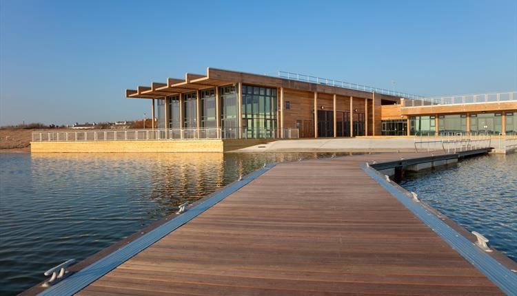 Modern wooden pavilion with glass walls and curved boardwalk extending over calm lake waters at sunset.