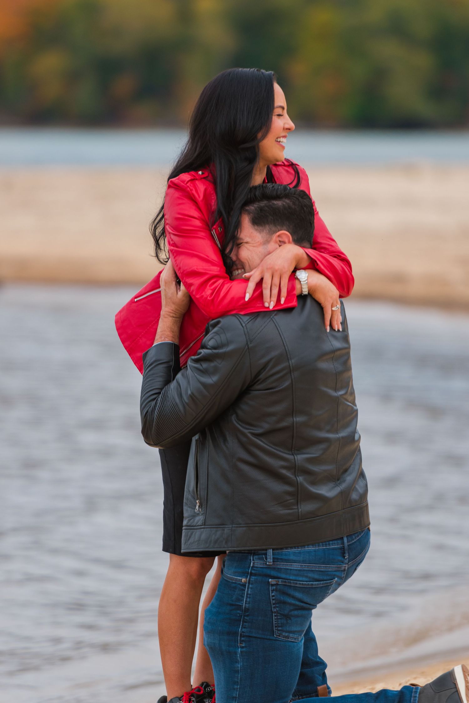 A couple embraces on a beach at sunset with their small beagle dog wearing a sweater sitting nearby.