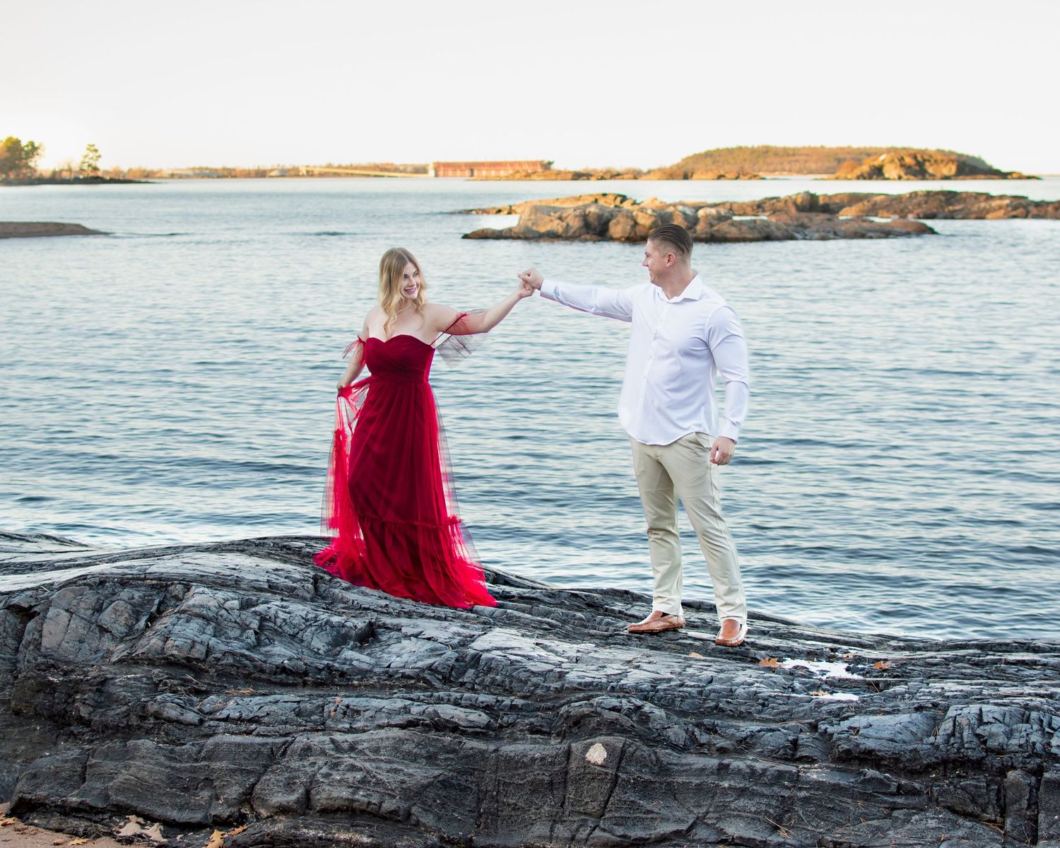 Couple in white shirt and red dress sitting on rocky shoreline during sunset engagement photoshoot.