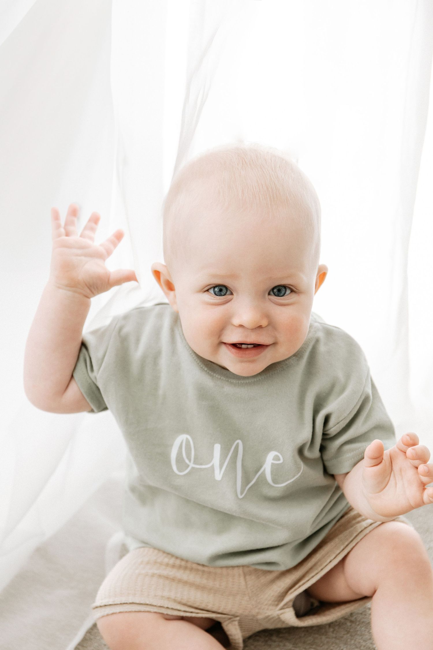 A smiling baby in a sage green "one" t-shirt with joyful expressions against a white background, during birthday shoot in Nottingham