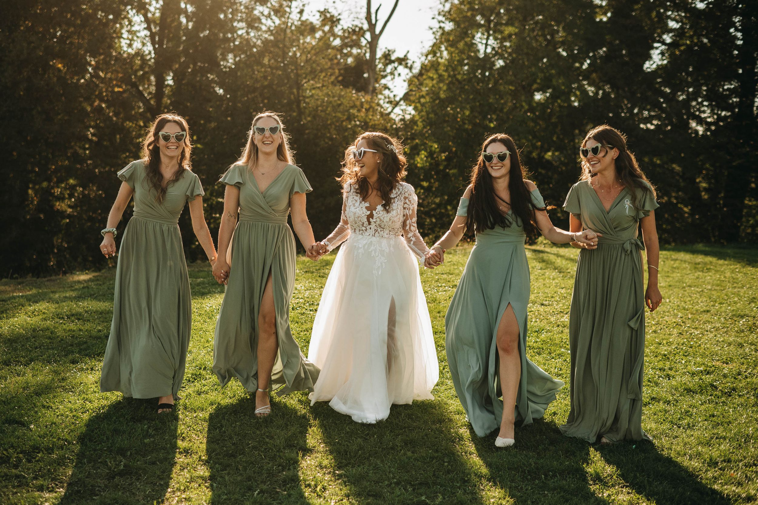 Bridesmaids in sage green dresses walking together on a sunny grassy hillside.