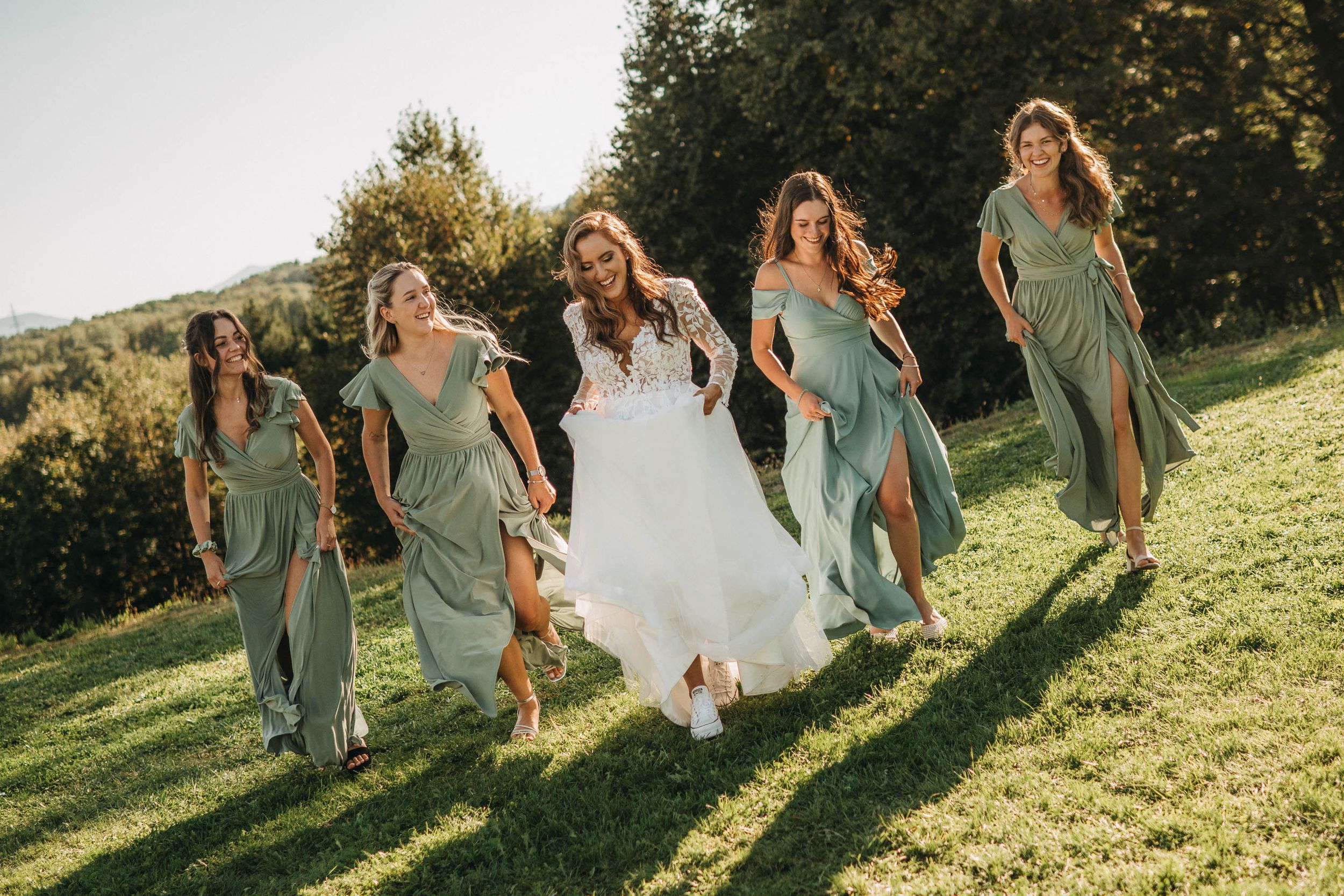 Bridesmaids in flowing sage dresses dancing together on a grassy slope.