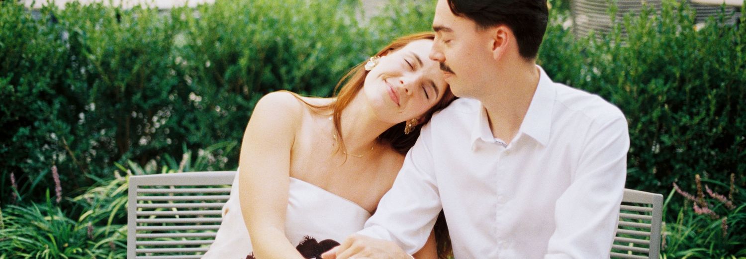 A couple shares a tender moment while sitting together on a white bench surrounded by lush greenery.