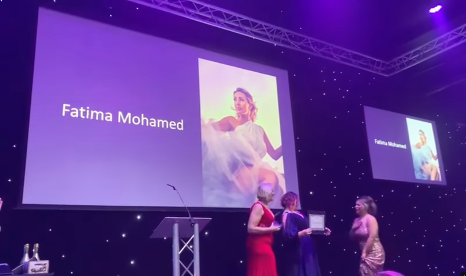 Conference presentation with purple stage lighting and multiple projection screens showing the winner Fatima Mohamed.