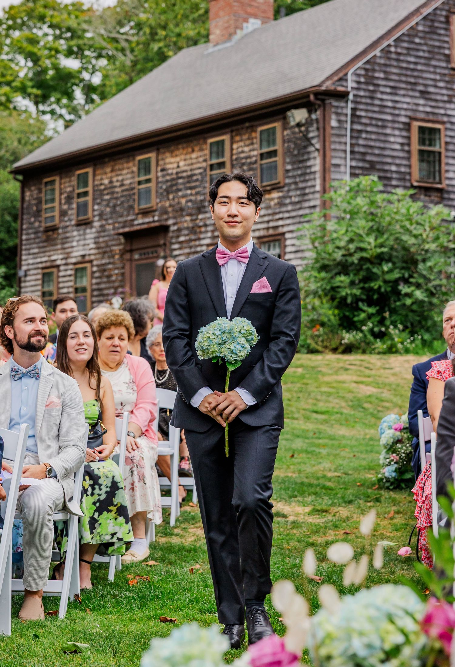Wedding guests seated on white chairs outdoors watch a ceremony unfold in front of a rustic wooden barn.