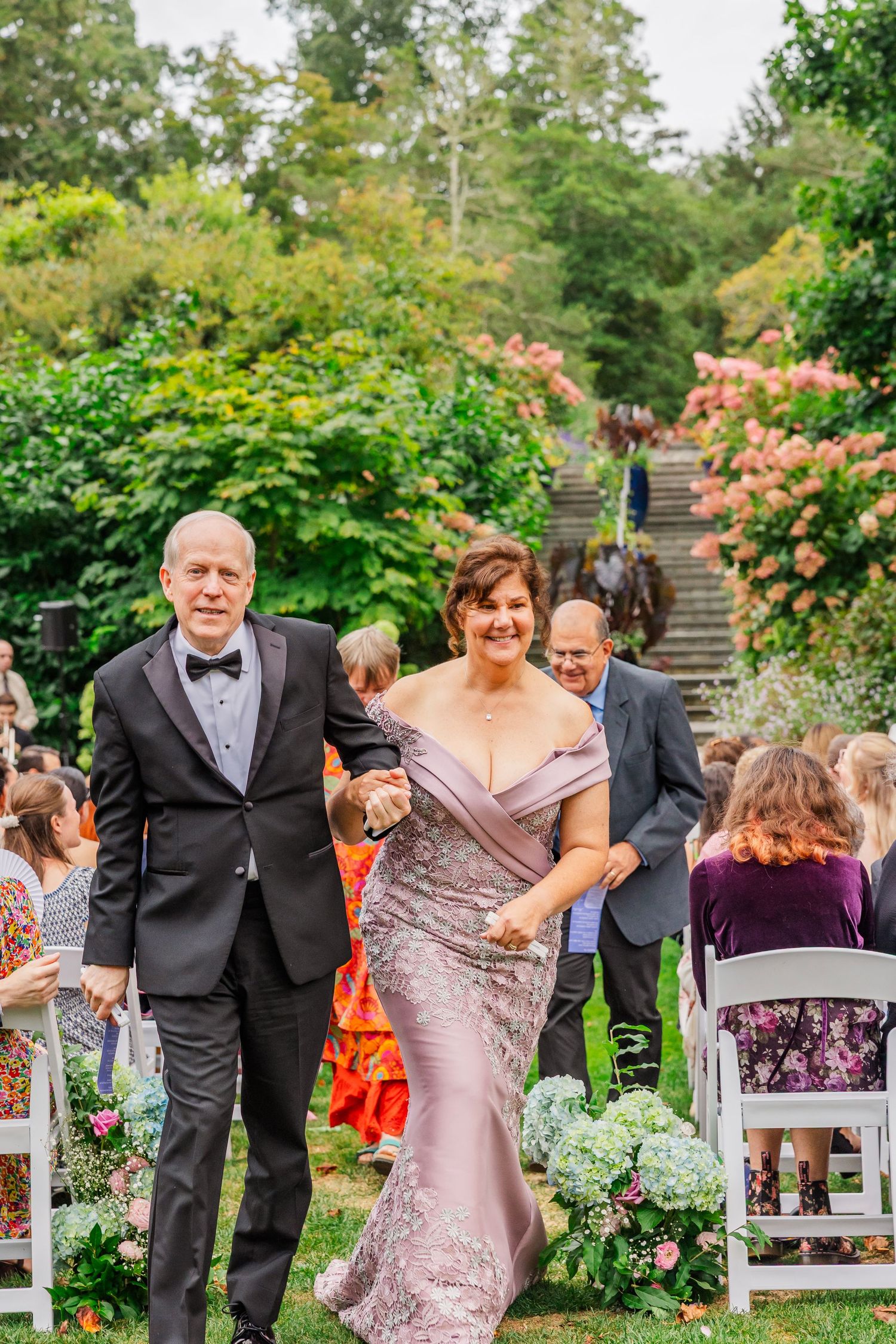 Wedding party walks down garden path surrounded by blooming roses and lush greenery at outdoor ceremony.