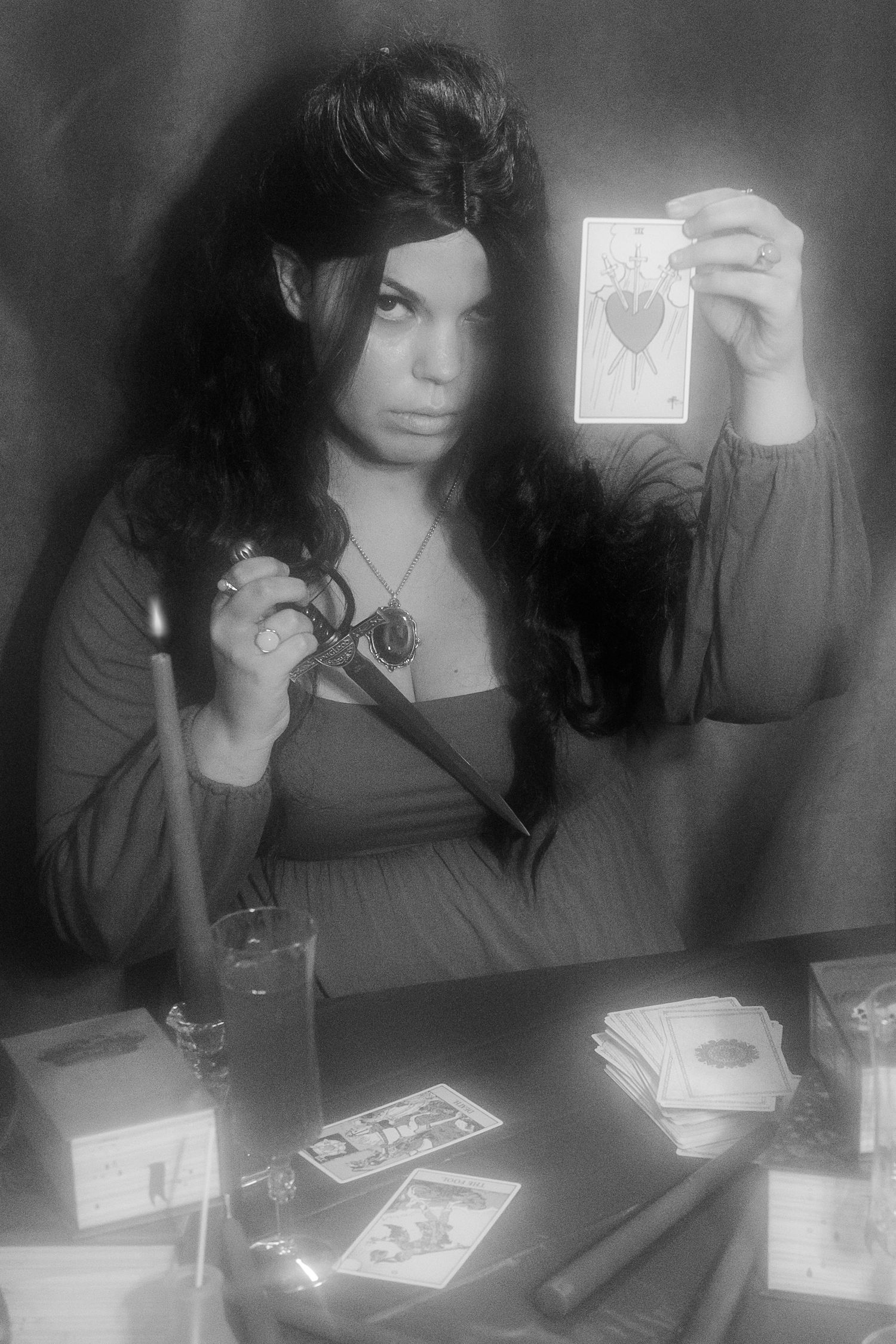 A black and white photograph shows a figure in dark clothing holding up a tarot card at a table with scattered cards.