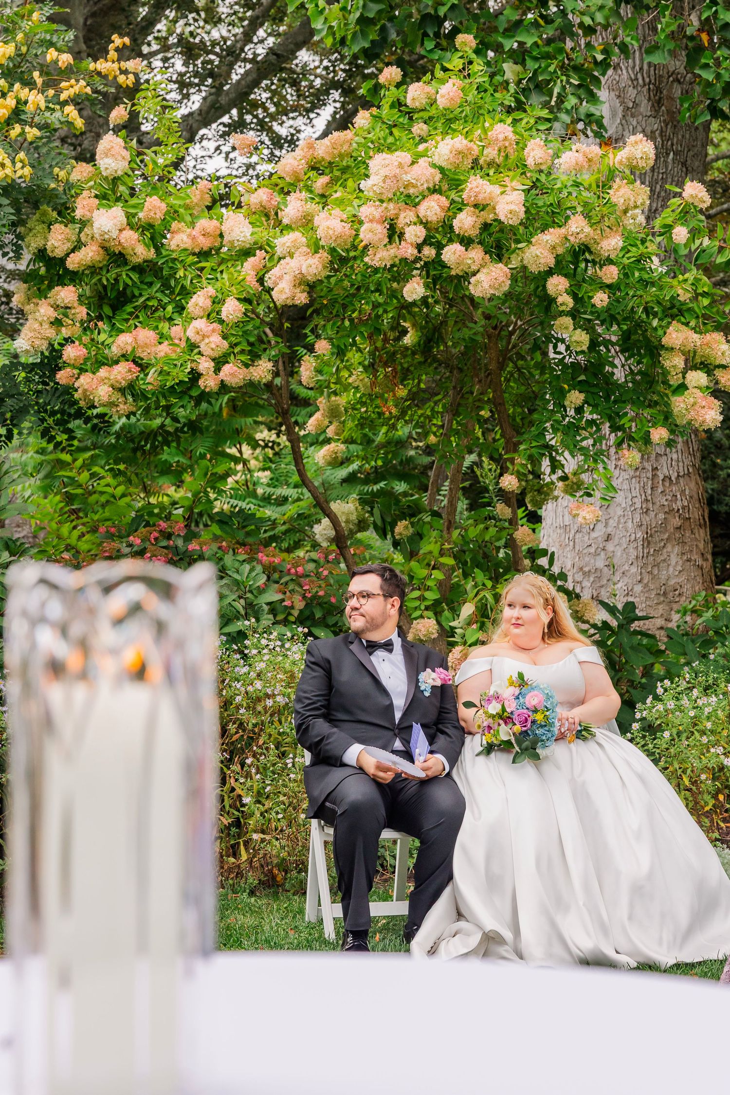 Couple sits on elegant white bench under blooming peach hydrangea trees in garden setting during wedding celebration.