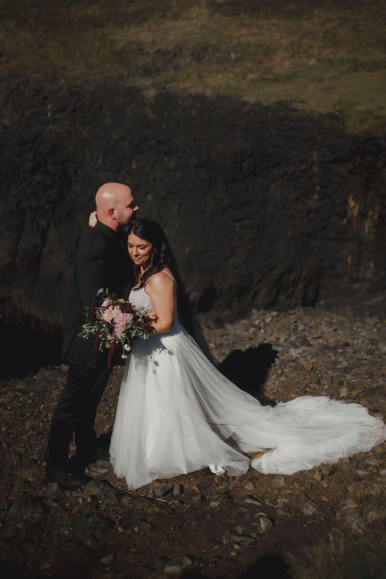 A romantic wedding photo of a couple embracing on a dark rocky backdrop with the bride holding a floral bouquet.
