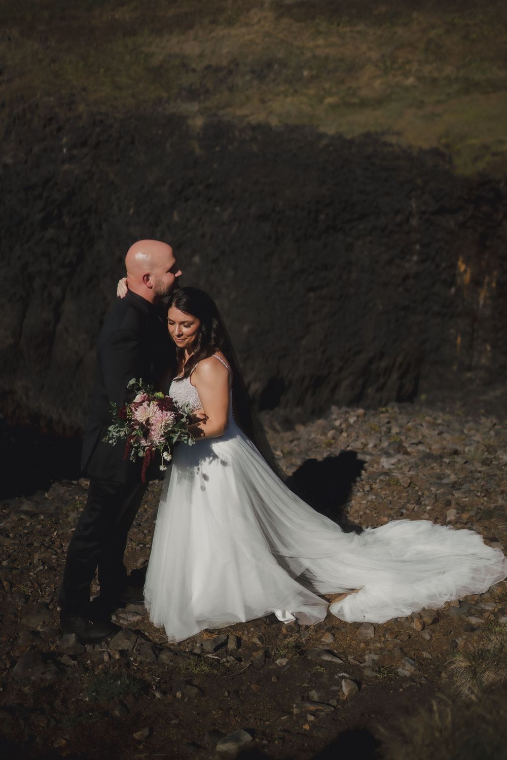 A romantic wedding photo of a couple embracing on a dark rocky backdrop with the bride holding a floral bouquet.