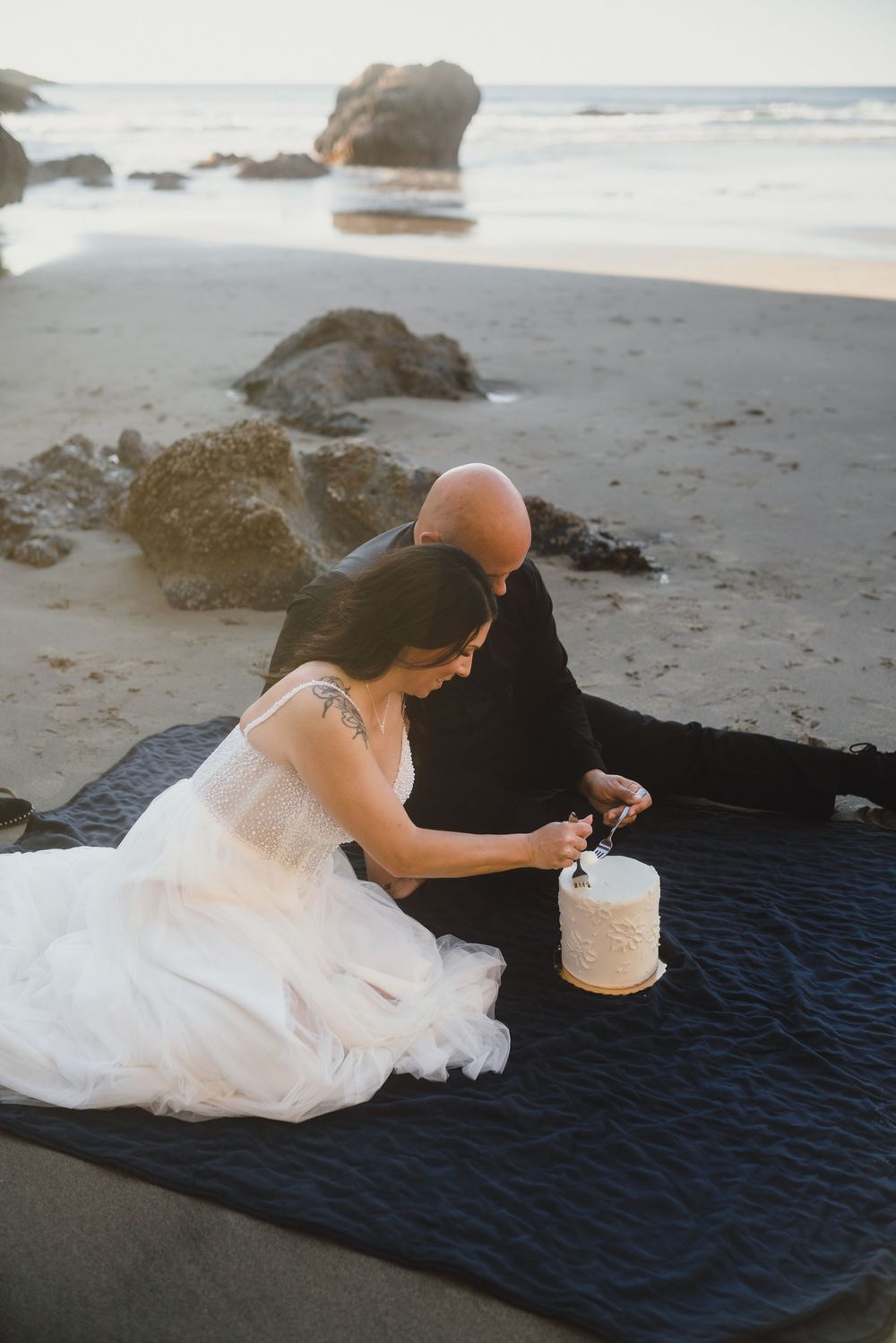 A couple sits on a blanket at the beach having a picnic in wedding attire with rocks and ocean in background.