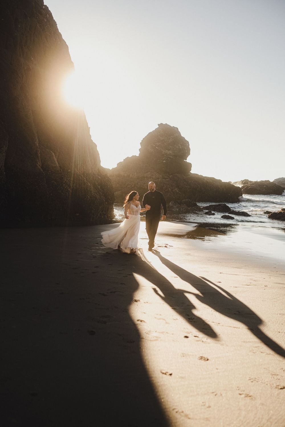 A couple casts long shadows on a sandy beach at sunset with towering coastal cliffs in the background.
