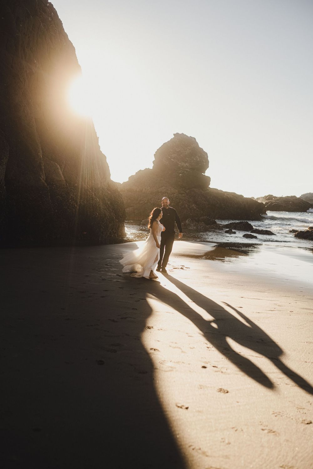 A silhouetted couple walks along a beach at sunset with dramatic cliffs and rocks casting long shadows.