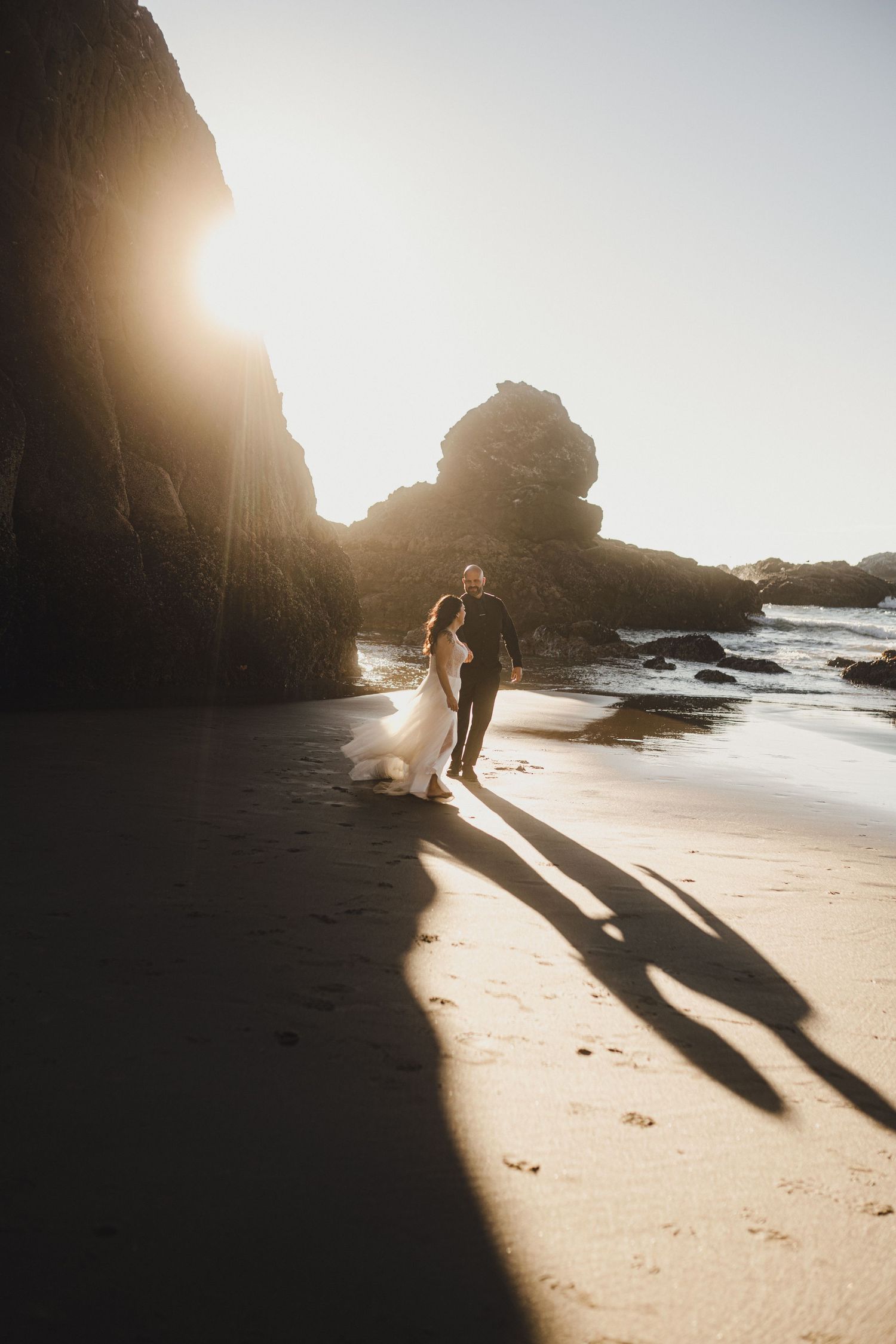 A silhouetted couple walks along a beach at sunset with dramatic cliffs and rocks casting long shadows.