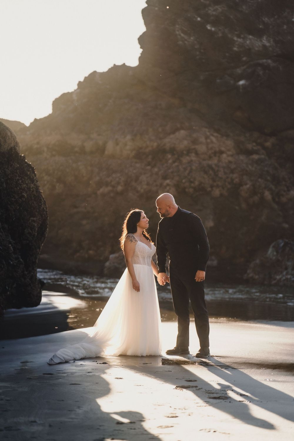A wedding couple shares an intimate moment on a beach at sunset with towering cliffs in the background.