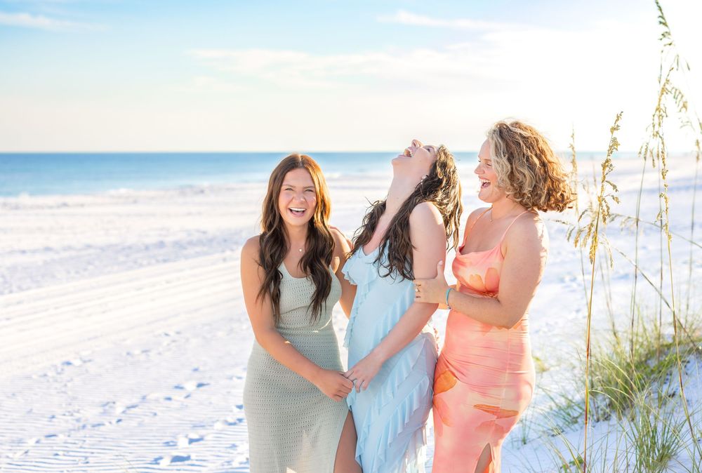 Three friends laugh together on a sunny beach with sea oats and white sand along the shoreline.