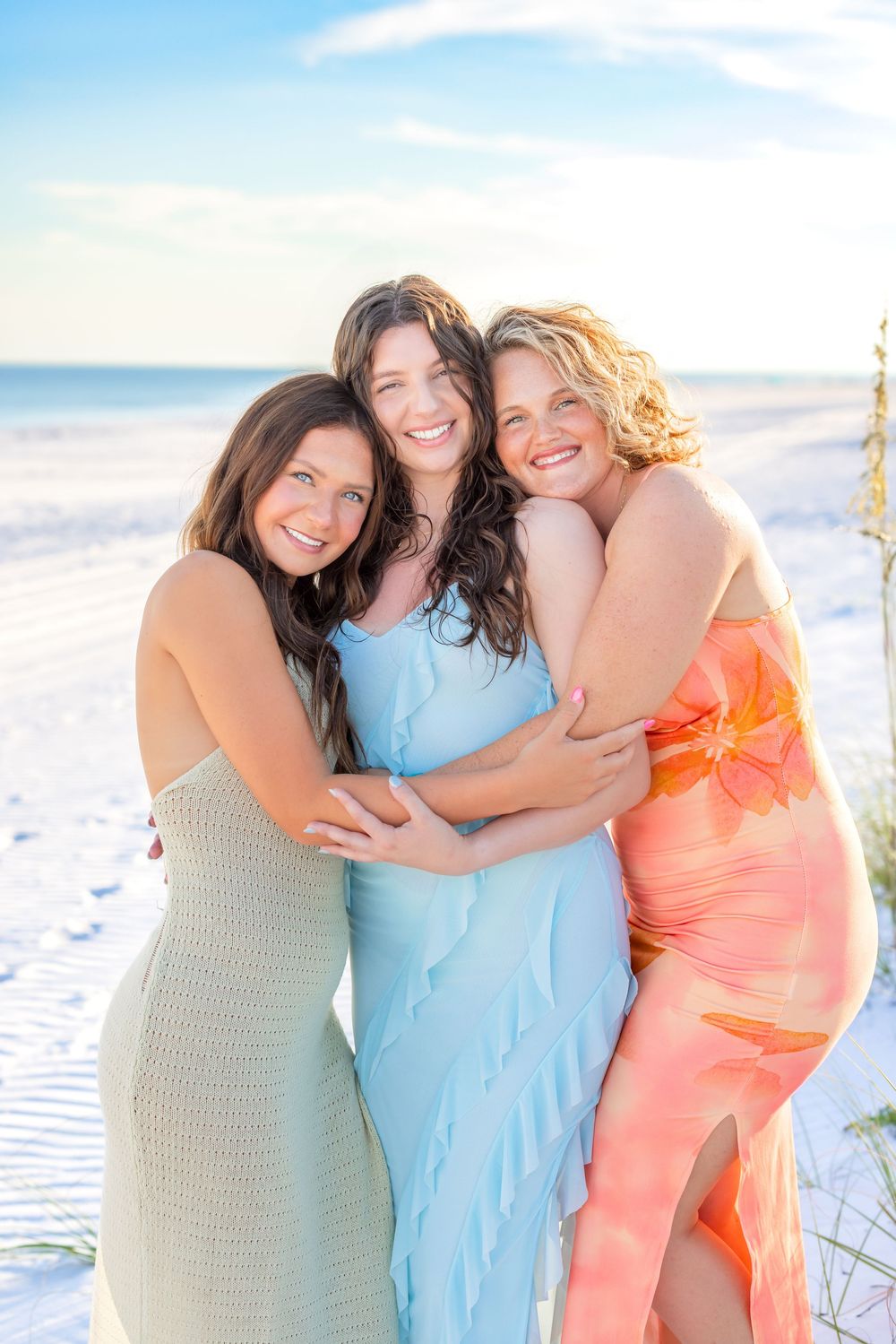 Three friends in pastel dresses embrace and laugh together on a sunny white sand beach at sunset.