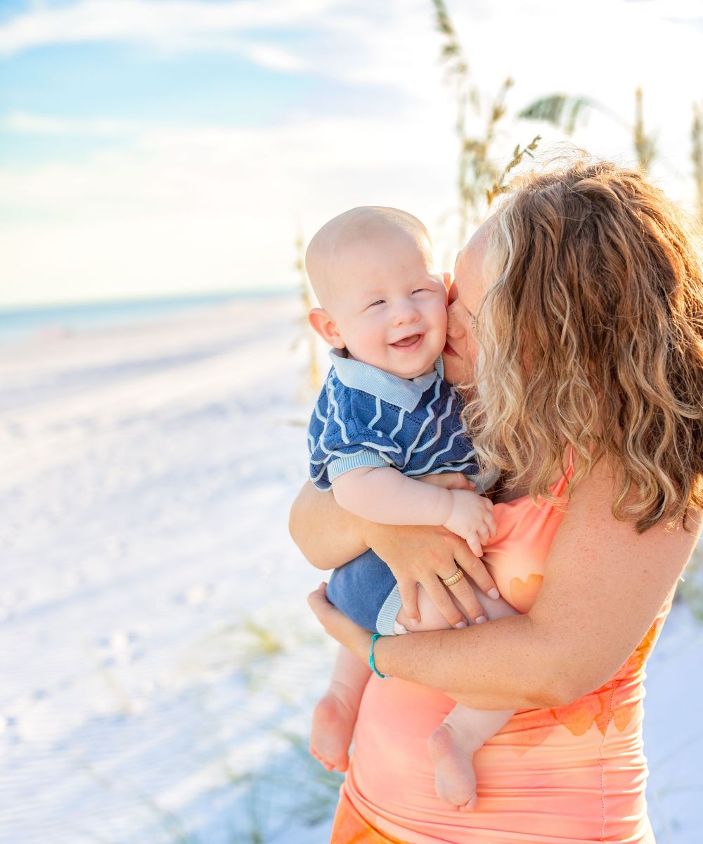 A parent in a coral top holds and embraces a small baby wearing blue against a bright beachy background.