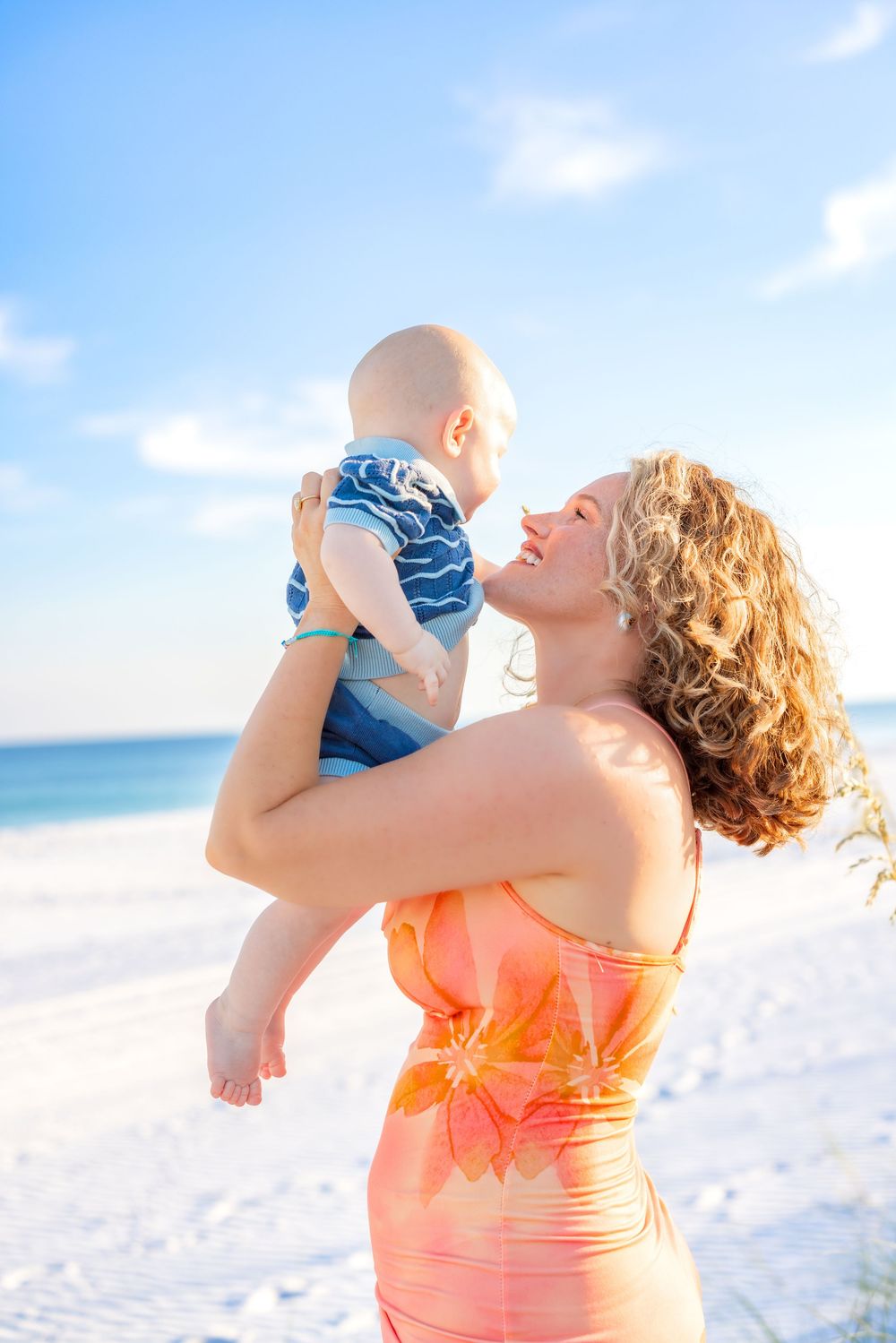 Woman in coral dress lifts baby wearing blue against white sandy beach and ocean backdrop.