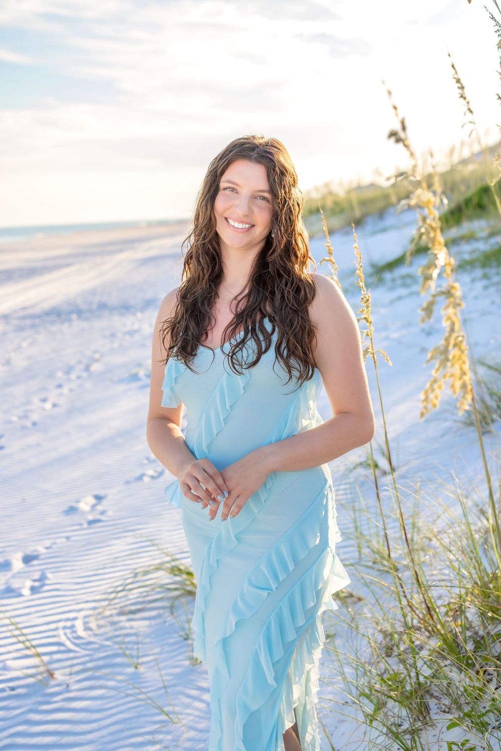 A person in a light blue dress poses on a sandy beach with sea oats and ocean waves during golden hour.