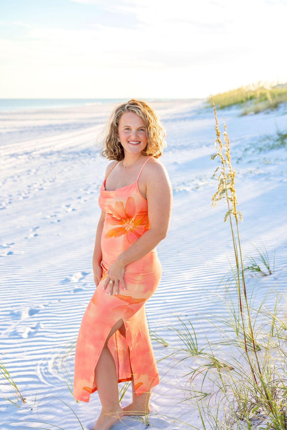 A person in a coral-colored dress poses on a sandy beach with sea oats and ocean waves in the background.
