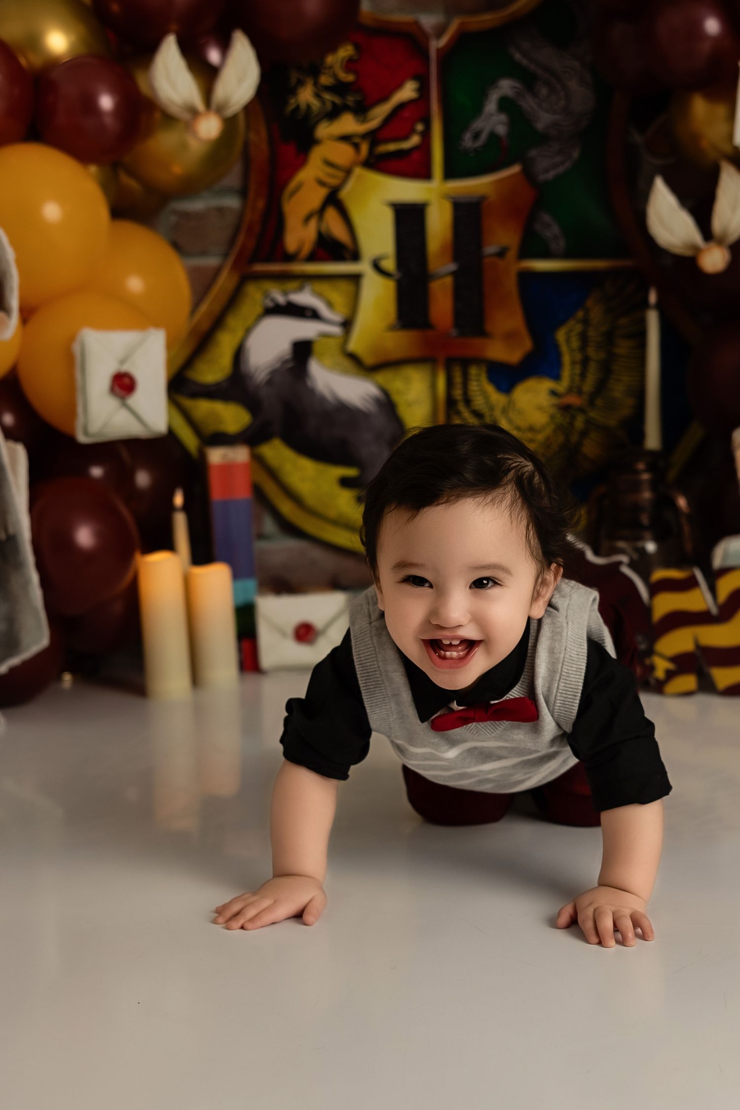 Adorable baby in formal bow tie attire crawls on bright white surface with colorful balloons and decorations in background.