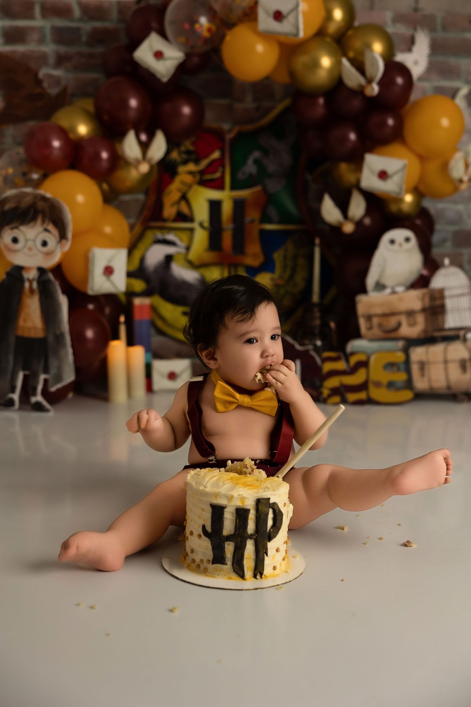 Harry Potter themed first birthday cake smash with maroon and gold balloon arch, magical decorations and props against brick wall.