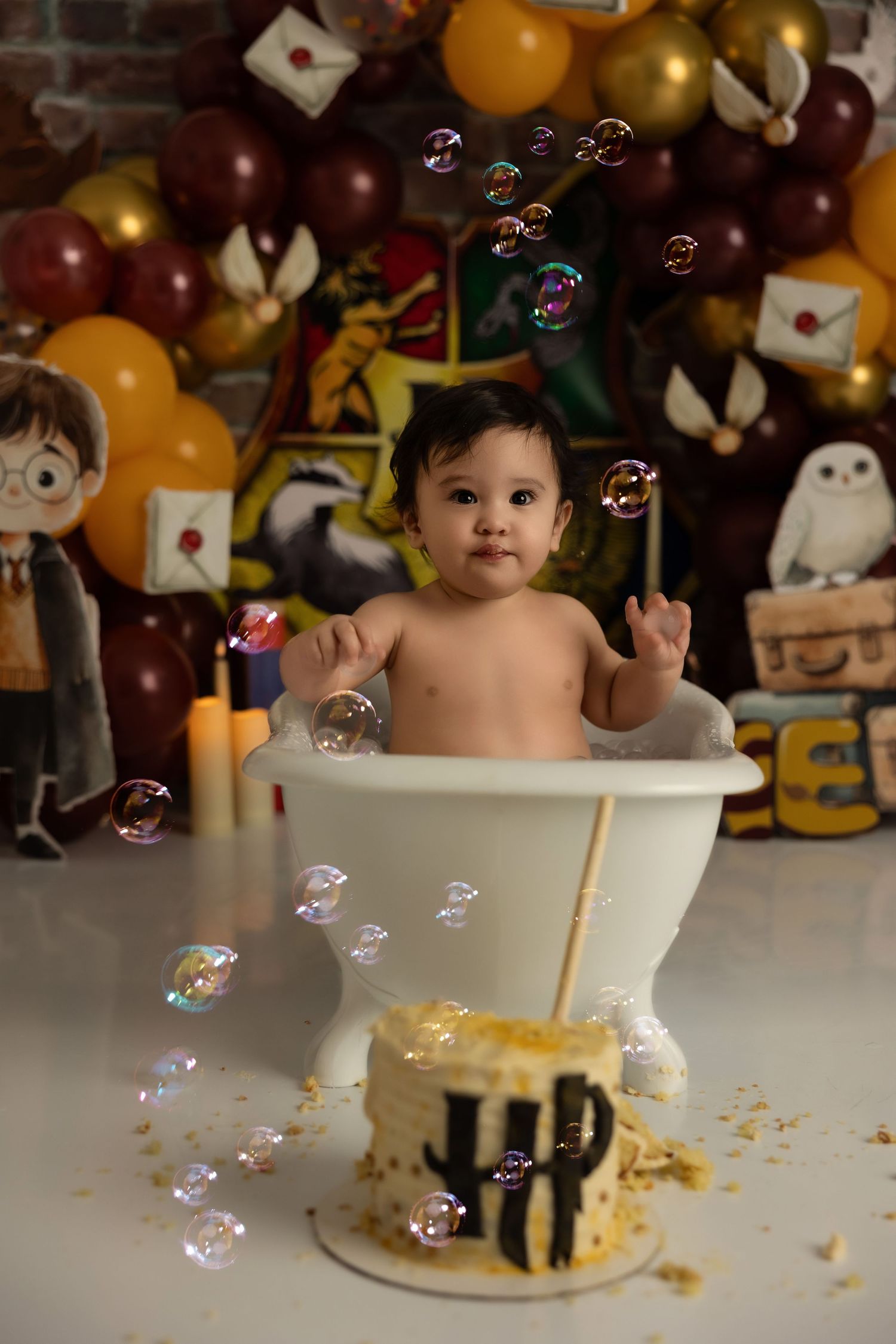 A festive balloon and stuffed animal display surrounds a bubble-filled cake smash photoshoot with decorated cake in foreground.