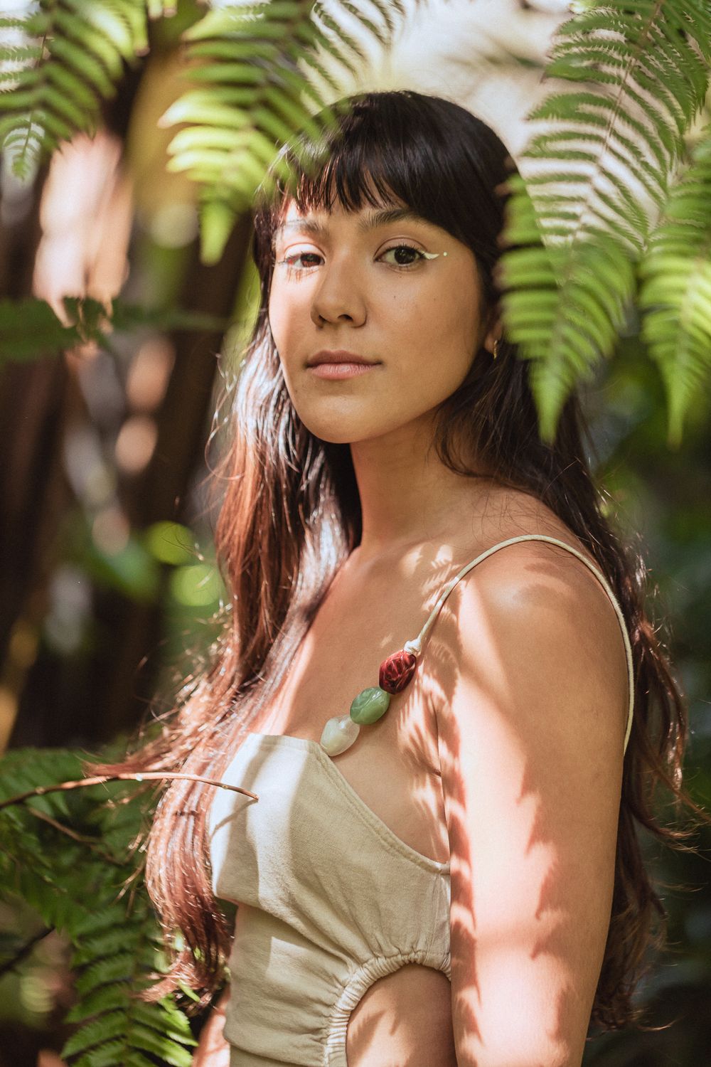 A portrait in dappled sunlight showing long dark hair and a beige dress framed by lush tropical ferns in the background.