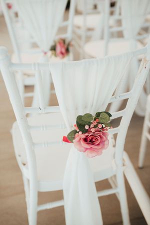 White wedding ceremony chair decorated with pink rose and draped white fabric for an elegant aisle setup.
