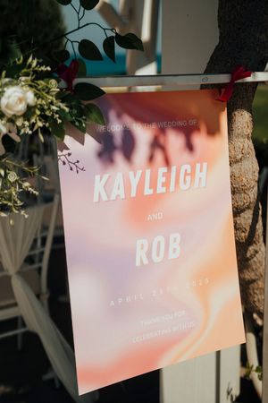A peach and pink gradient wedding invitation for Kayleigh and Rob hangs on display with floral decorations nearby.