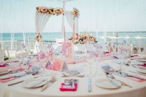Long banquet tables decorated in soft pink and white settings with floral centerpieces and ocean views through glass windows.