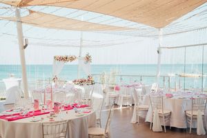 Elegant beachfront wedding reception setup with white chairs, pink table accents and flowing fabric canopy overlooking the ocean.
