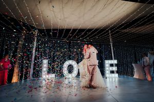 A couple shares their first dance at a wedding reception with glowing LOVE letters and falling confetti on the dance floor.